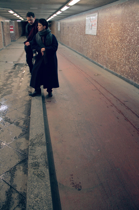 Zwei Personen stehen einem Tunnel. Beide Personen sind weiß und tragen dunkle Wintermäntel und Schals. Die vordere Person sieht nach links, die hintere Person sieht auf den Boden.