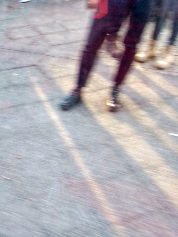 Die Farbfotografie zeigt die Beine von drei Personen. Alle Personen tragen enge Hose und zwei der Personen tragen goldene Boots. Das Sonnenlicht fällt in Streifen auf die Straße.