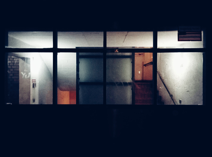 A photograph. Shot from the street through a large window displaying a dull lit ground floor with dirty white walls of an old building. One staircase is leading up to the next floor, the little lighted space at the top is lit in a grey dull orange light. One staircase is leading down to the basement, the space there is lit in dark orange. An elevator shaft is built in the middle. Its opaque green glas front turns the dim lights inside the elevator into a dark green. In the left corner there's an entrance to a dark hallway with grey brick walls.