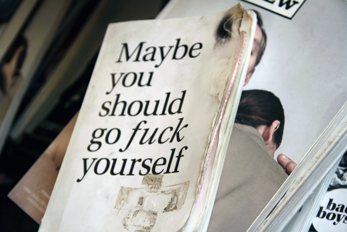 Ein Buch mit verkohlten Seiten und einem Brandfleck liegt auf einem Magazin. In schwarzen Buchstaben steht auf dem weißen Buchcover": "Maybe you should go fuck yourself." Das Titelbild des darunterliegenden Magazins zeigt zwei Menschen, das Gesicht des einen Menschen ist durch das verbrannte Buchcover verdeckt, das Gesicht des anderen Menschen sieht von der Kamera weg.